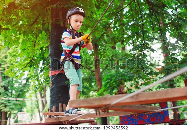 The boy climbs a rope park. Little cute boy in a\
climbing adventure park. Summer sunny day. Rope playground\
structure. Safe climbing extreme sport. Child with helmet insurance\
in a rope park.