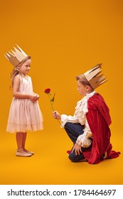 A Boy In Costume Of Prince And Gives A Rose To A Girl In A Princess Costume. Childhood Dreams. Fantasy, Imagination. Studio Portrait On A Yellow Background.