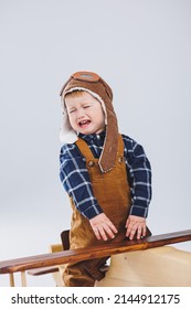 The Boy Is Crying In A Leather Jacket And A Pilot's Hat, A Wooden Plane, Brown Suitcases. Children's Wooden Toys. Baby Emotions