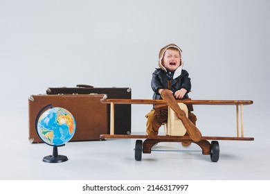 The Boy Is Crying In A Leather Jacket And A Pilot's Hat, A Wooden Plane, Brown Suitcases. Children's Wooden Toys. Baby Emotions