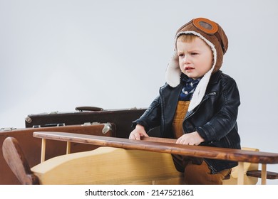 The Boy Is Crying In A Leather Jacket And A Pilot's Hat, A Wooden Plane, Brown Suitcases. Children's Wooden Toys. Baby Emotions