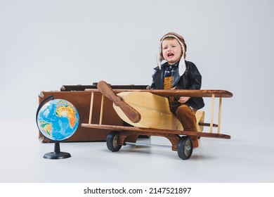 The Boy Is Crying In A Leather Jacket And A Pilot's Hat, A Wooden Plane, Brown Suitcases. Children's Wooden Toys. Baby Emotions