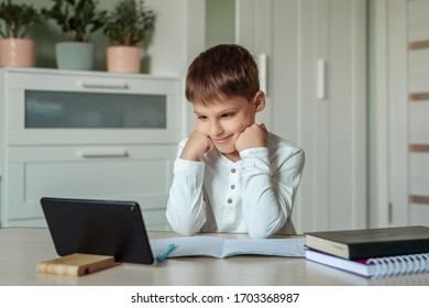 A Boy With Dark Hair In A White Shirt Sits At A Table And Does Homework Using A Tablet. Distance Learning At Home, Doing Homework During The Epidemic And Pandemic Covid-19