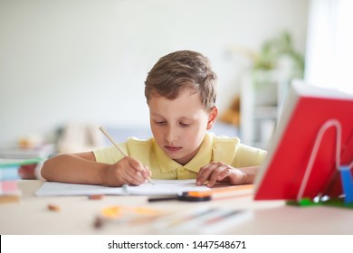 The Boy Does His Homework At Home. Happy Child At The Table With School Supplies Concentrated Writing In The Retreat, Doing Homework . Positive Student In A Bright Room With A Pencil In His Hands.