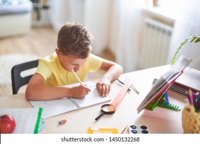 The Boy Does His Homework At Home. Happy Child At The Table With School Supplies Concentrated Writing In The Retreat, Doing Homework. Positive Student In A Bright Room With A Pencil In His Hands.