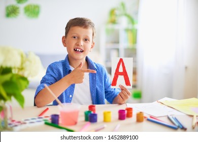 boy does his homework at home. a happy child at the table with school supplies smiles funny and learns the alphabet in a playful way.positive student in a bright room with painted letters in his hands - Shutterstock ID 1452889100