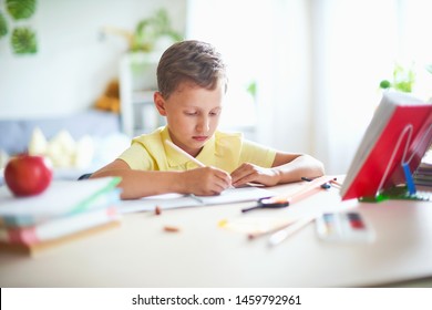 The Boy Does His Homework At Home. Happy Child At The Table With School Supplies Concentrated Writing In The Retreat, Doing Homework. Positive Student In A Bright Room With A Pencil In His Hands.