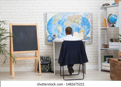 The Boy Does His Homework At Home. Happy Child At The Table With School Supplies Concentrated Writing In The Retreat, Doing Homework. Positive Student In A Bright Room. A View From The Back