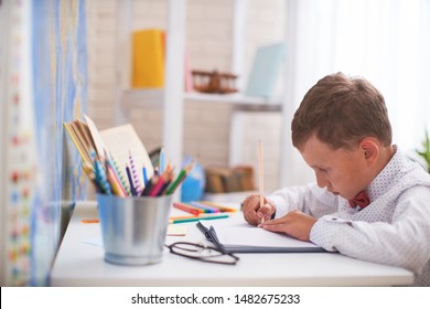 The Boy Does His Homework At Home. Happy Child At The Table With School Supplies Concentrated Writing In The Retreat, Doing Homework . Positive Student In A Bright Room With A Pencil In His Hands.