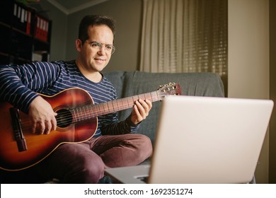 Boy Doing An Online Course To Learn How To Play The Classical Guitar. He Is Sitting On The Sofa At Home, Playing The Guitar In Front Of The Laptop While Listening To The Lessons