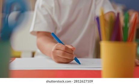 Boy drawing with blue crayon, hand on paper, sketching a face near a yellow pencil holder on red table in studio; creative concentration.