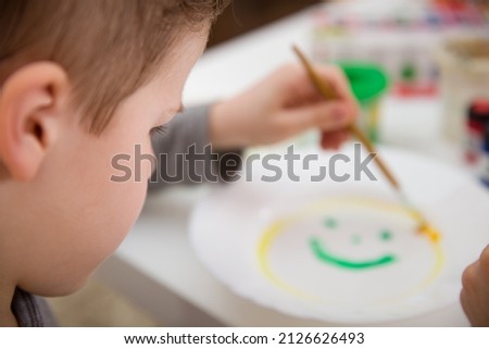 The boy draws with a brush and paints on a white plate sitting at the table. Hobbies, activities for children. Early development. Creative gifts