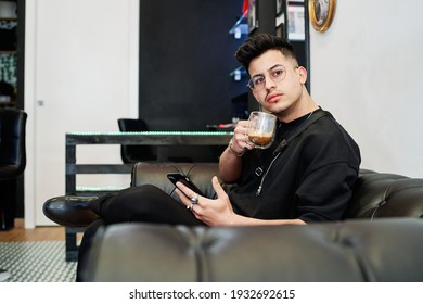 boy dressed in black sits drinking coffee while waiting for his turn in a barber shop - Powered by Shutterstock