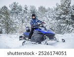 Boy driving snowmobile in a winter landscape. Little boy on the snowmobile up in the mountains smiling in a snowy weather, forest. Child rides a blue snowmobile on a snowy trail, pine trees.