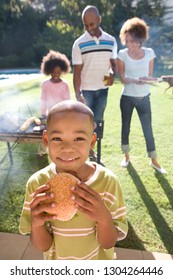 Boy Eating Burger Family At Barbeque In Garden At Camera