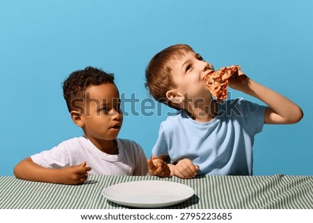 Boy eating pizza slice while friend watches beside empty plate on blue background. Concept of playful pizza sharing, kids snack break, food envy, and fast food campaign.