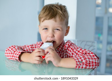 Boy eats a chocolate bar while sitting in kitchen. Guy sits at the table and eats a chocolate bar. 