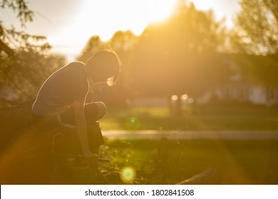 Boy Exploring Quietly Outside In The Evening Light, Sitting Thoughtfully As The Sun Sets. Beautiful Sunflare And Backlighting On Boy. 