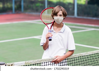 Boy In Face Mask Playing Tennis On Outdoor Court During Coronavirus Outbreak. Teenager With Tennis Racket And Ball In Sport Club. Active Exercise For Kids. Social Distancing. Training In Pandemic. 