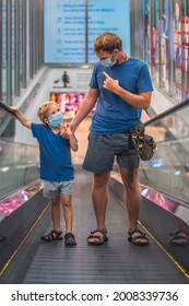 Boy Father Going Up Super Market Escalator, Wear Protect Mask, COVID Coronavirus Pandemic Lockdown Quarantine. Dad Hold Child Hand Looking At Kid Finger Point Show Face. New Life Family Shopping VERT