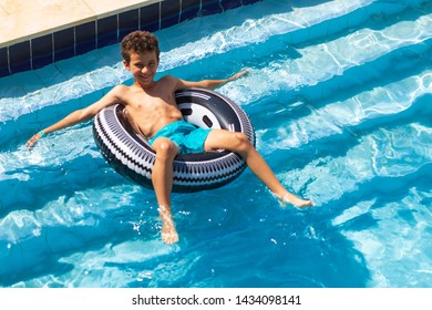 Boy Floating On An Inflatable Black Circle In The Pool. Closes Eyes From Bright Sunshine