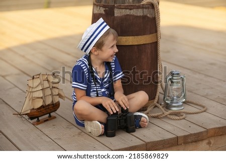 A boy in the form of a sailor sits on a wooden deck near a brown barrel. Model of a sailing ship. Kerosene lamp. Summer season, warm sunset lighting.