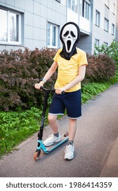 Boy In Ghostface Mask From The Scream Movie Riding A Scooter On Asphalt Path Near House Wall And Bushes - Saint Petersburg, Russia, June 2021