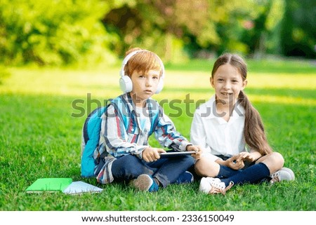 a boy and a girl with backpacks are sitting on the lawn and reading a book and a tablet with headphones, the concept is back to school, brother and sister are doing homework on the street
