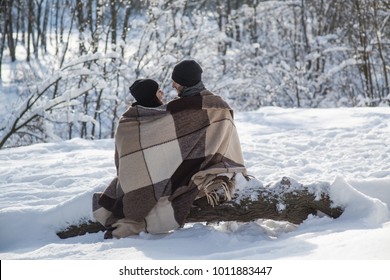 Boy And Girl In Black Hats And Plaid Sitting On The Snow Background. Couple Sitting On A Log.