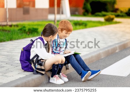 a boy and a girl with briefcases or backpacks are sitting at the school and reading a book and tablet, going back to school, brother and sister are studying, doing homework on the street