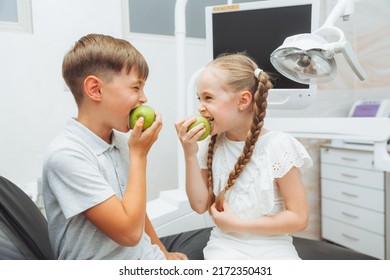 A Boy And A Girl Are Eating Apples In The Dentist's Chair. The Concept Of Healthy Teeth. Children Eat A Green Apple.