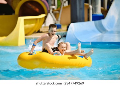 Boy and girl have fun on water slide in outdoor aquapark. Little children floating on yellow inflatable raft.