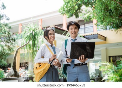 Boy And Girl Of Indonesian High School Students Smiling At The Camera As They Stand Holding Book, Computer Laptop, And Wearing Bag