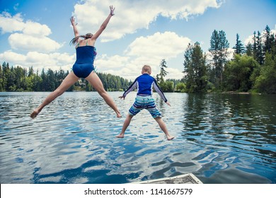 Boy And Girl Jumping Off The Dock Into A Beautiful Mountain Lake. Having Fun On A Summer Vacation. View From Behind