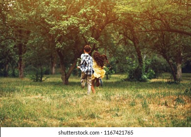 Boy And Girl Running In Distance Holding Hands. Romantic Little Couple Of Sweet Children Running Away Into Nature Holding Hands Together.