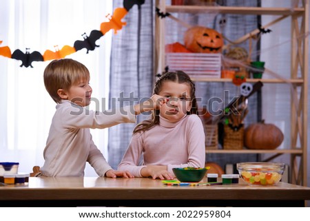 Boy and girl sit at a table with treats and pumpkins in a room decorated for Halloween. The boy teases his sister with jelly worms. Halloween. Close up.