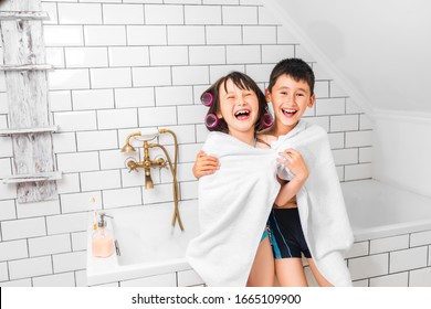 Boy And Girl Sitting On The Edge Of The Bathroom, Wrapped In One Towel. The Kids Have Fun Laughing. Next To A Toothbrush And A Bottle Of Liquid Soap.