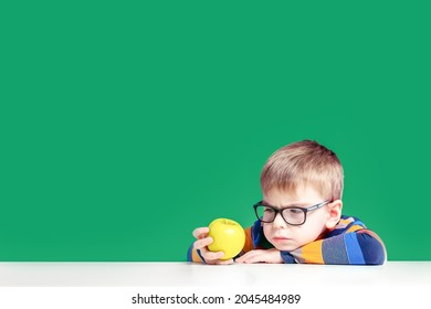 The boy in glasses frowning pensively looks at the apple he is holding in his hand.
