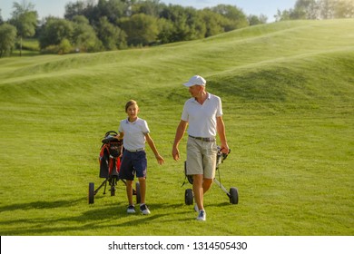The boy golfer with his father or trainer pulling their carts on perfect golf course at summer evening