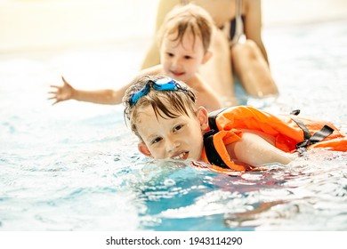 Boy With Googles And Safety Vest Learns To Swim Near Brother And Mom In Large Pool With Clear Water At Sports Complex Closeup