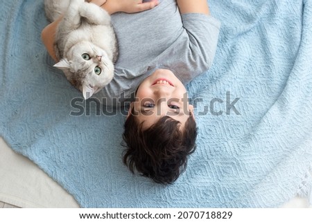 A boy in a gray T-shirt lies on the floor on a knitted blue plaid, with a beautiful white cat, smile