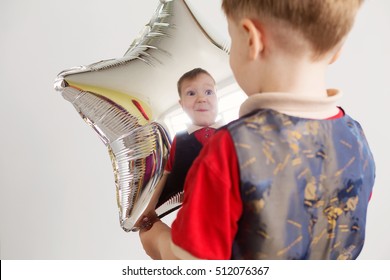 Boy Grimacing And Playing The Ape With Star-shaped Balloons In Studio. Kid Looks And Rejoices At His Reflection In Foil Balloon. Child Laughing Looking At The Reflection In A Distorted Mirror