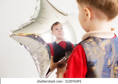 Boy Grimacing And Playing The Ape With Star-shaped Balloons In Studio. Kid Looks And Rejoices At His Reflection In Foil Balloon. Child Laughing Looking At The Reflection In A Distorted Mirror