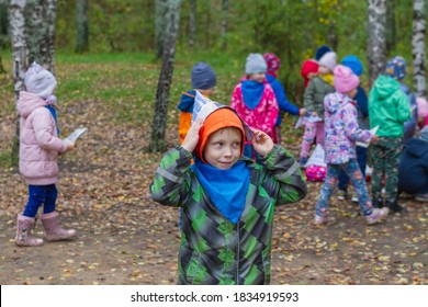 The Boy Has A Newspaper Hat On His Head. Funny Boy Child In An Orange Hoodie And A Green Jacket Smiling. A Child Walks On The Playground.