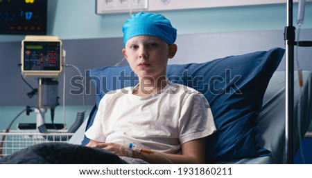 Boy in hat looking at camera while lying on bed in ward of modern cancer hospital