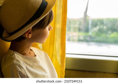 A boy in a hat looks out the window thoughtfully while sitting on a tour bus. 