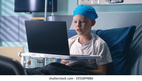 Niño en el sombrero saludando a la mano y hablando mientras hace videollamadas en una laptop en el hospital de oncología