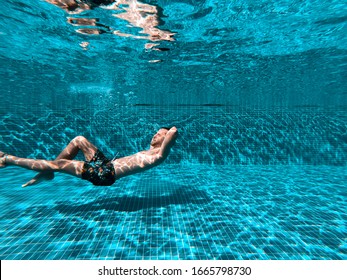 A boy in a Hawaiian-style swimsuit is swimming underwater with his arms crossed at the back of his neck. Beautiful shadows and reflections of the sun in the pool. Lifestyle water. Fun in the pool. - Powered by Shutterstock