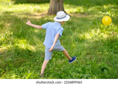 A boy in a headdress, shorts, shirt plays with a yellow ball on the lawn on a sunny summer day.The concept of vacation, vacation, active sports. Copy space. - Shutterstock ID 2256899365