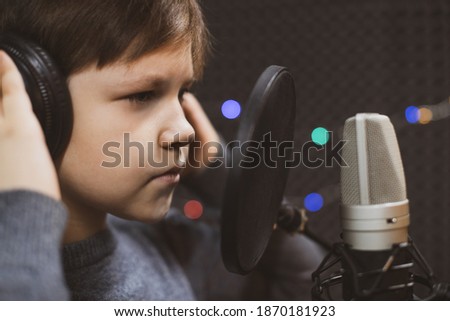 Boy in headphone is singing or talking into microphone with pop filter in voice recording studio. Young singer recording his voice for phonogram performance for entertainment.